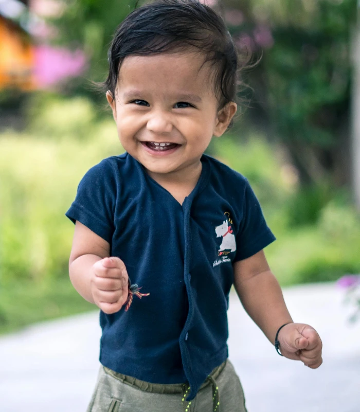 A happy toddler grinning and standing on a sunny outdoor path with greenery.