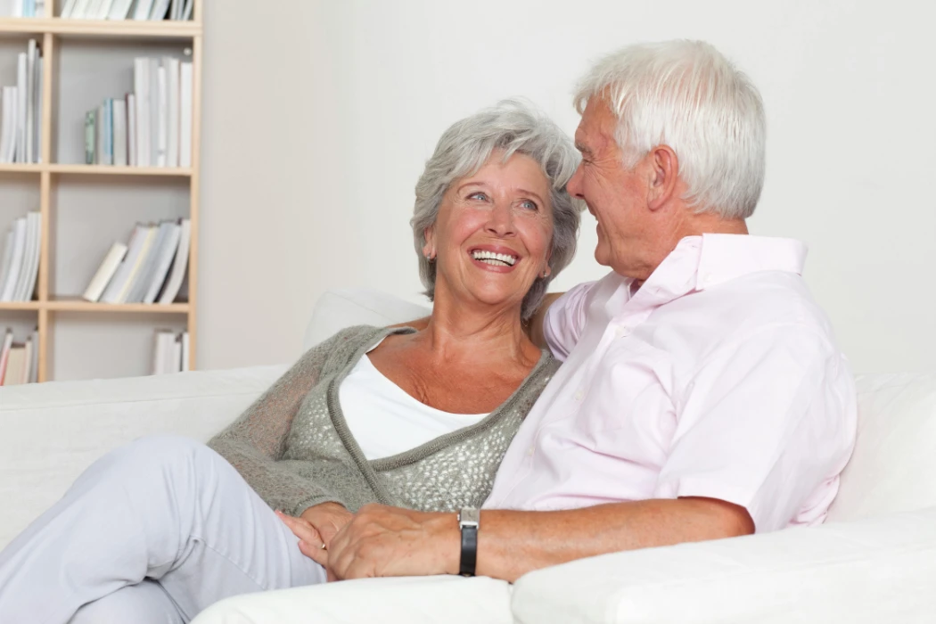Smiling senior Caucasian couple sitting comfortably on a sofa in a bright room full of books.